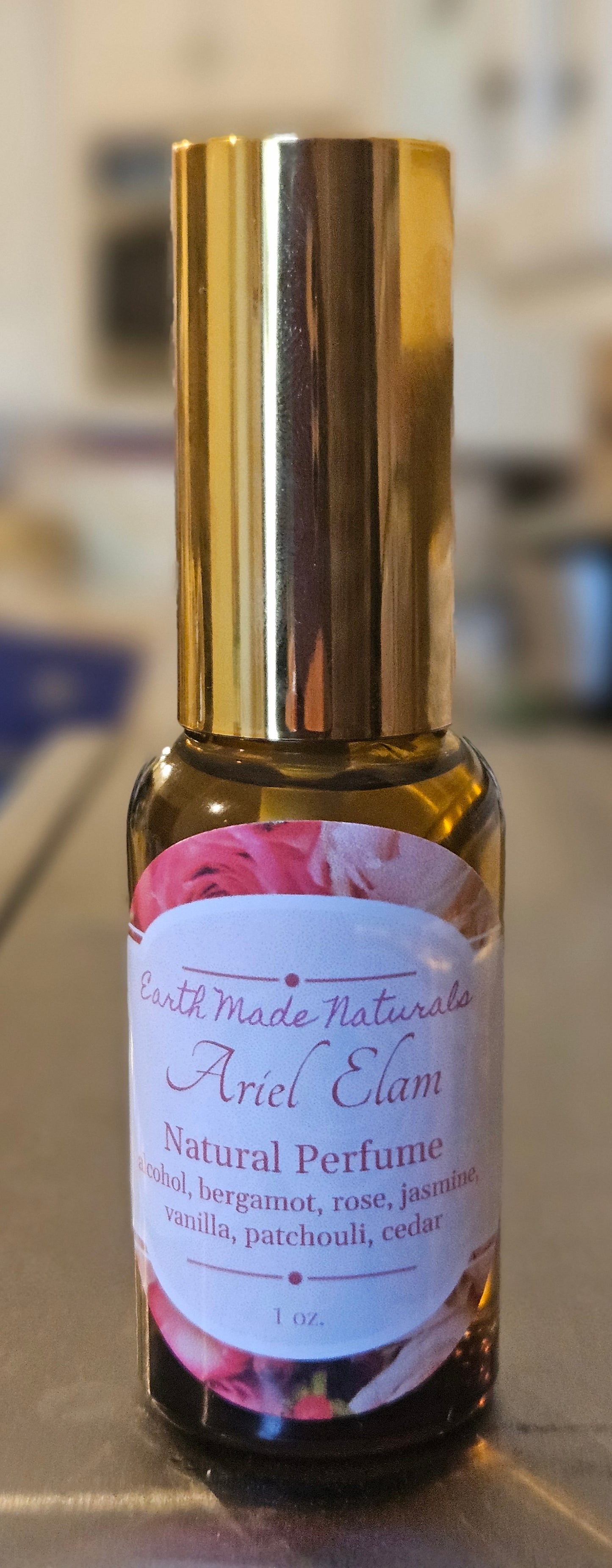 A small glass bottle of natural perfume with a shiny gold cap and a white label featuring pink floral artwork. The label reads 'South Made Naturals Ariel Elam Natural Perfume'.