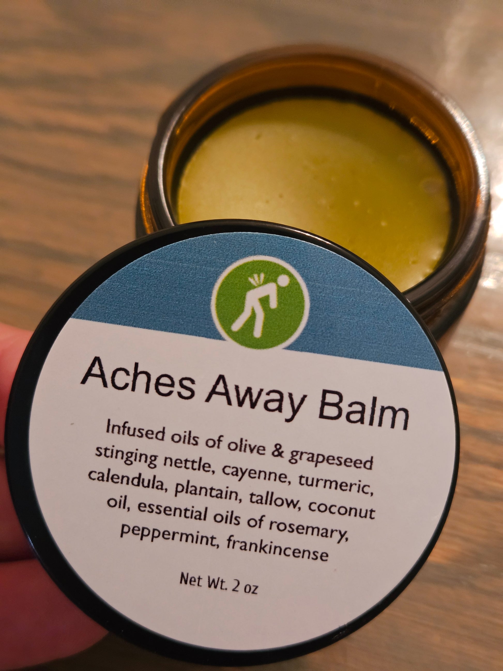 A close-up of a hand holding the lid of an amber glass jar labeled 'Aches Away Balm,' with the open jar showing a pale yellow balm behind it on a wooden surface.