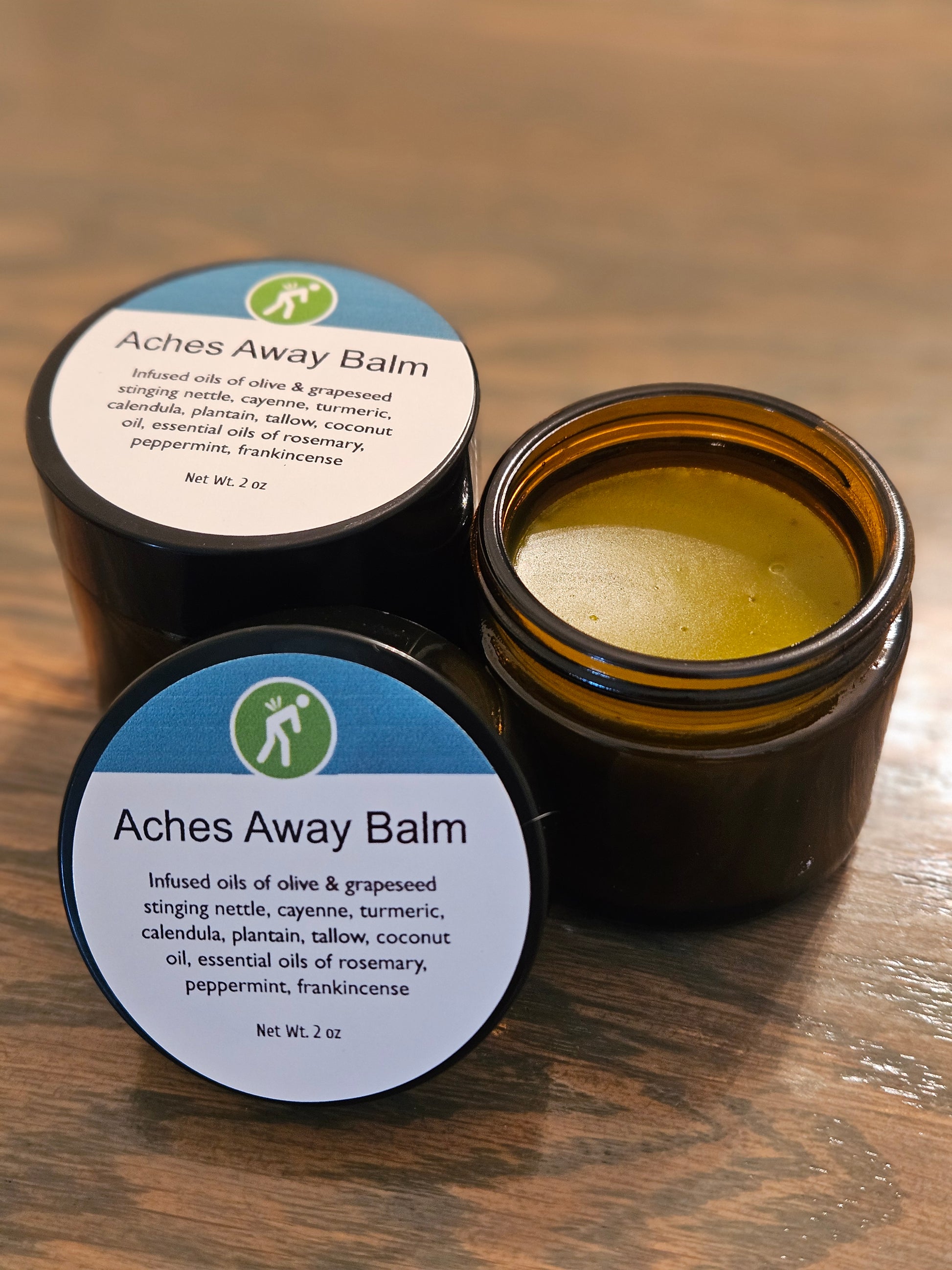Two amber glass jars of Aches Away Balm on a wooden surface, with one jar open showing the yellow balm inside and the other showing the closed lid with the product label.