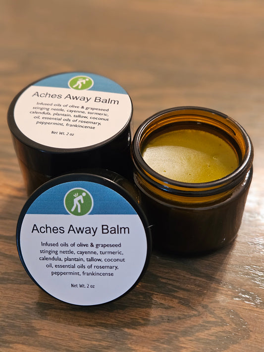 Two amber glass jars of Aches Away Balm on a wooden surface, with one jar open showing the yellow balm inside and the other showing the closed lid with the product label.