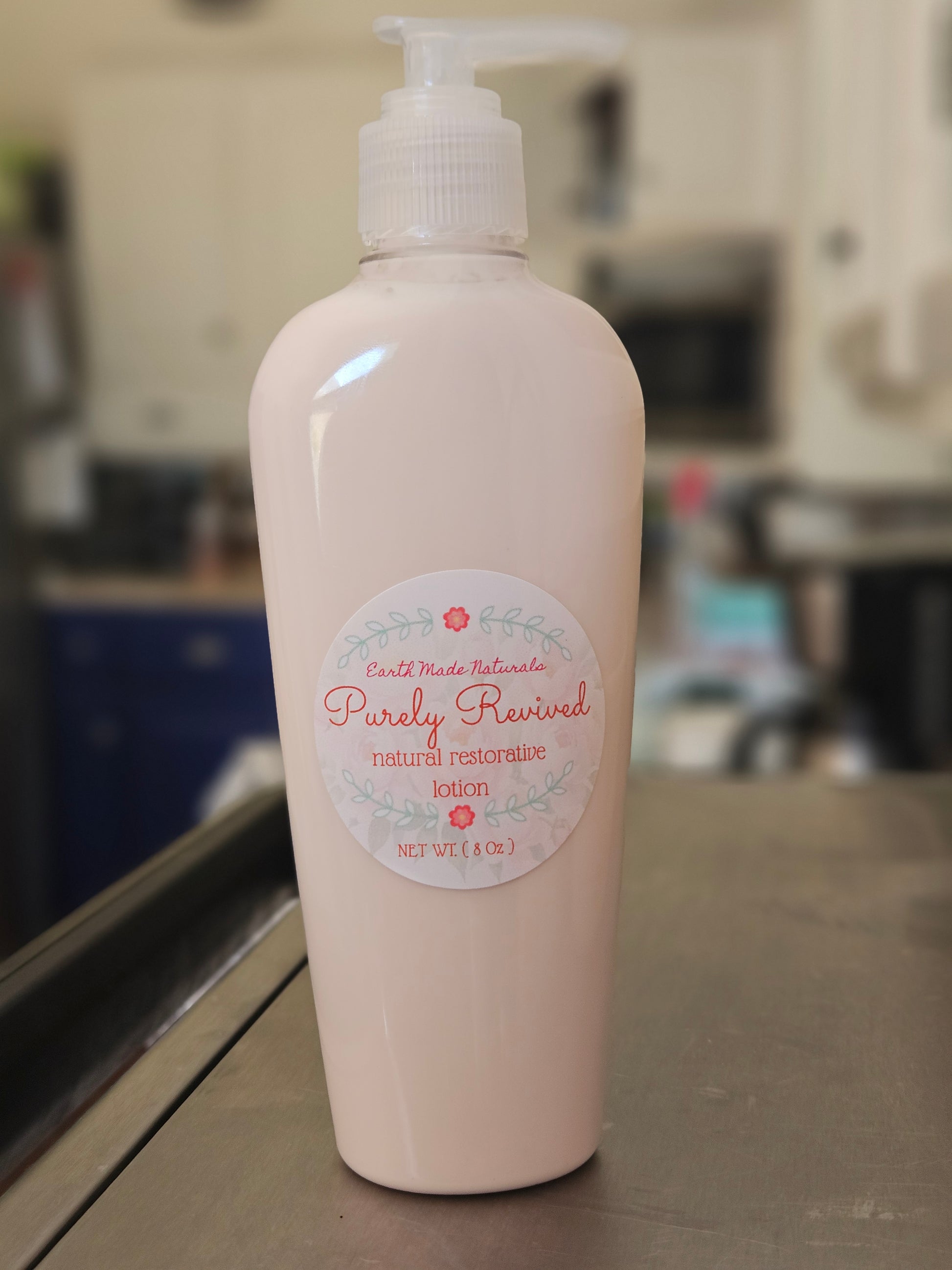 A bottle of Earth Made Naturalists Purely Revived Natural Moisturizing Lotion in a pale pink color, standing on a countertop with a blurred kitchen background.