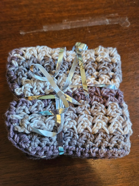 handmade crocheted dish cloths in a striped pattern of beige, gray, and mauve-purple, wrapped in clear cellophane and tied with a shiny silver ribbon, set on a dark wooden surface.