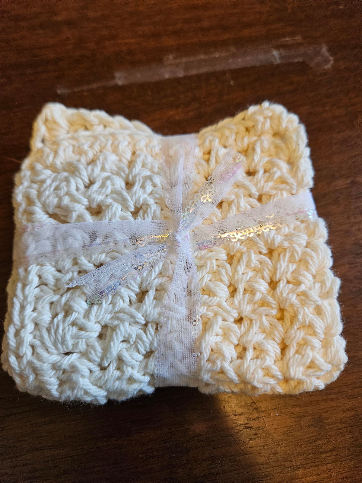 A folded cream-colored crocheted textile item, likely a baby blanket or swaddle, wrapped with a sheer white ribbon and a small gold sequin bow, placed on a dark wooden surface.