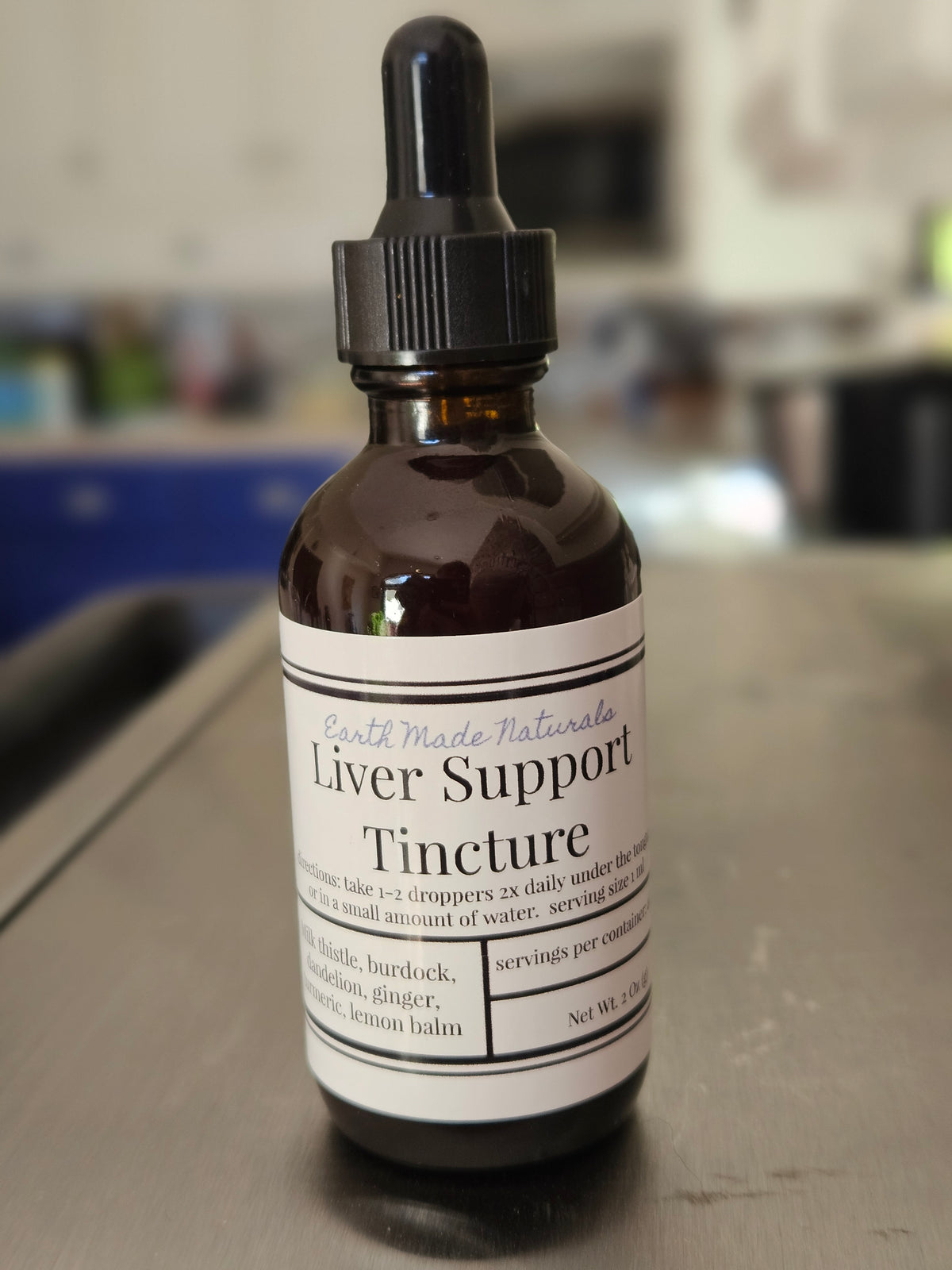 An amber glass dropper bottle of Earth Made Naturals Liver Support Tincture, sitting on a dark tabletop with a blurred indoor background.