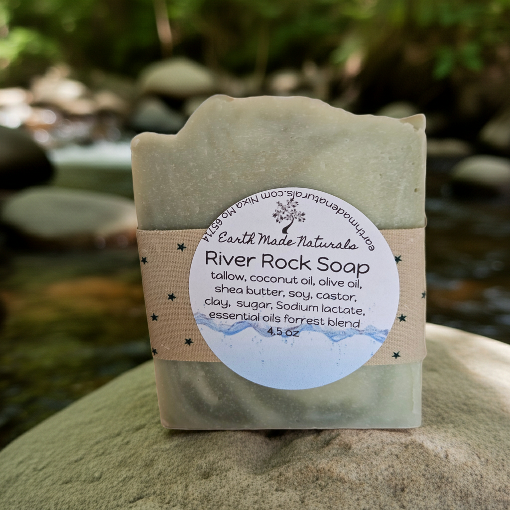 A bar of Earth Made Naturals River Rock Soap is wrapped in a kraft paper band with a round white label, set against a blurred outdoor background of a stream and rocks.
