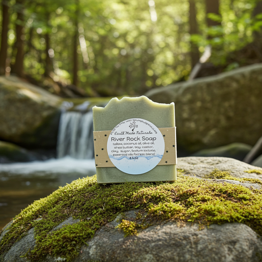 A bar of River Rock Soap by Craft Made Naturals is displayed on a mossy rock in a forest setting with a blurred waterfall in the background.