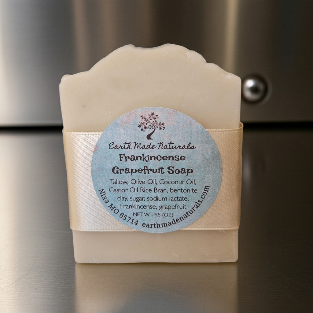 A bar of Earth Made Naturals Frankincense Grapefruit Soap, wrapped in a cream-colored paper band with a circular light blue label, set against a dark metallic background.