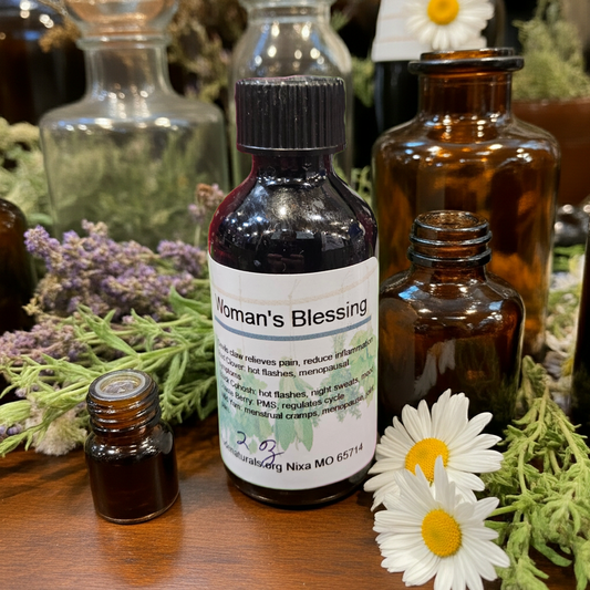 Bottle labeled 'Woman's Blessing' surrounded by herbs and flowers on a wooden surface