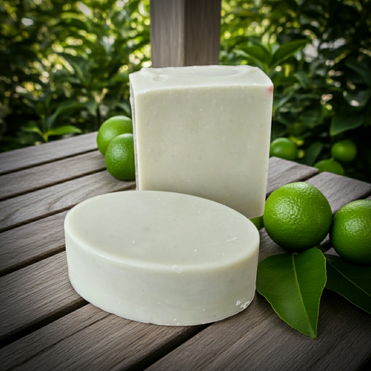 Two blocks of white soap on a wooden surface with green limes and leaves in the background.