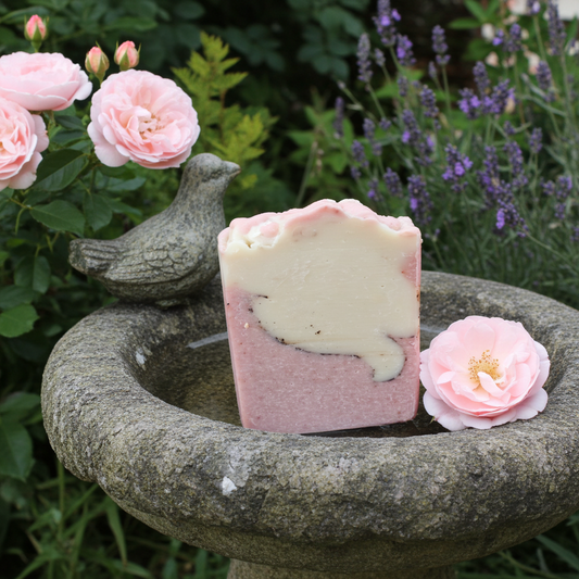 Bar of English Garden soap with pink and white gradient on a stone surface surrounded by flowers and a bird sculpture.